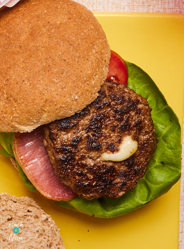 A close-up shows a Melt-in-the-Middle Bacon Cheeseburger , served in a bun with lettuce and tomato. The burger is well browned on the outside, and a bubble of melted cheese is oozing out of the centre.