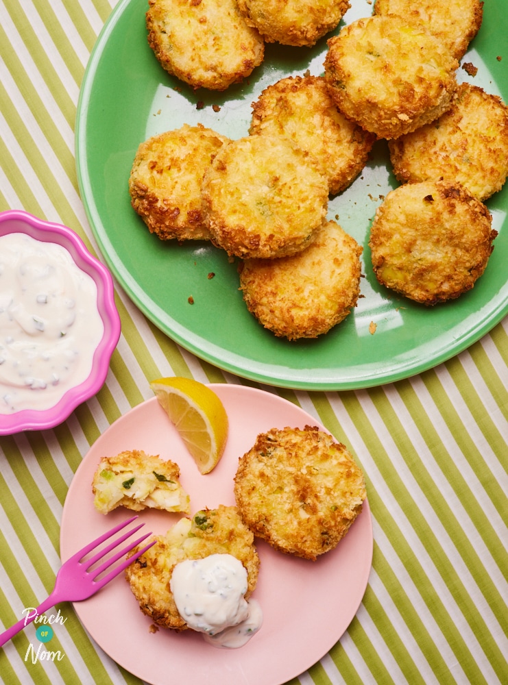 A table is set with a plateful of Pinch of Nom's slimming-friendly Smoked Haddock and Spring Onion Fishcakes, with one fishcake served on a smaller pink plate. A pink fork is cutting into the fishcake, revealing the mashed potato centre. There is a lemon wedge to the side to squeeze on top, and a homemade dip in a small bowl.