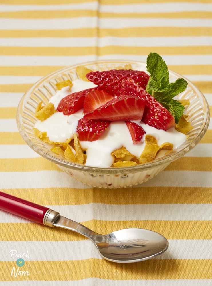 A bowl of Pinch of Nom's Summer Berry Dessert is served on a colourful, summery table topped with sliced strawberries and a creamy yoghurt with a spoon waiting to dig in.