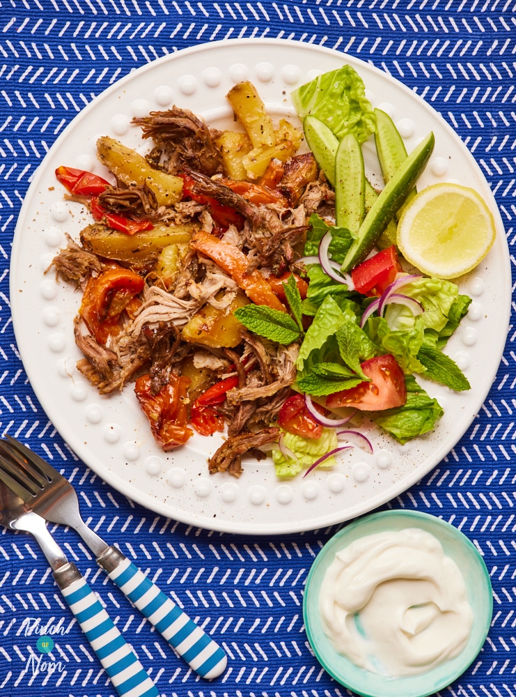 A table is set with a colourful plate of Pinch of Nom's slimming-friendly Kleftiko Lamb. The shredded lamb is served with a crispy, fresh side salad and a small bowl of sauce.