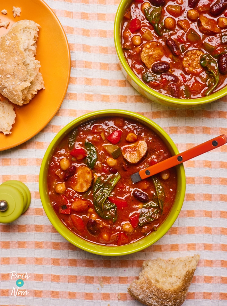 A table is set with two hearty bowls of Pinch of Nom's slimming-friendly Slow Cooker Chunky Tomato Cajun-Style Soup in bright yellow bowls, a crusty wholemeal bread roll waits to one side for mopping up every last drop.