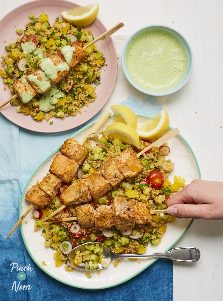 A hand reaches for one of Pinch of Nom's slimming-friendly Spicy Salmon Skewers with Couscous, served at the centre of a summery table with a garnish of lemon wedges and extra yoghurt drizzle on the side.