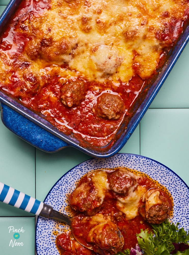 Pinch of Nom's slimming-friendly Baked Spicy Meatballs recipe is served in a large casserole dish, fresh from the oven at the centre of a blue worktop. A serving has been plated up to one side with a fork waiting to tuck in. The bake is topped with a bubbly layer of browned mozzarella and Parmesan.