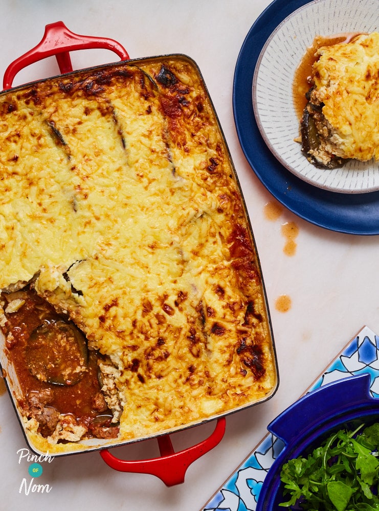 A large oven dish is filled with Pinch of Nom's Lamb Moussaka. It has a golden brown, cheesy topping and a meaty filling with lots of gravy. A large spoonful has been removed from one corner of the oven dish and served on a small plate.