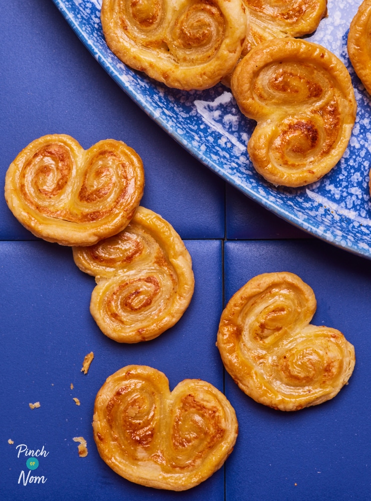 Pinch of Nom's slimming-friendly Cheese Palmiers are served scatted over a dark blue worktop and serving plate, looking golden brown and crisp.