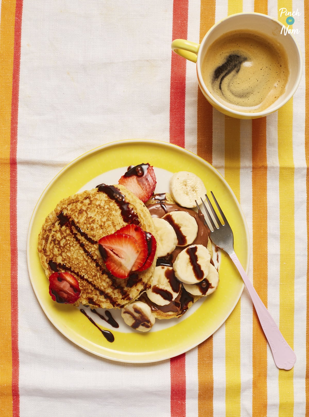 Two pancakes are served on a bright yellow plate, they're covered in banana slices, strawberries and drizzled with chocolate next to a frothy coffee in a yellow mug.