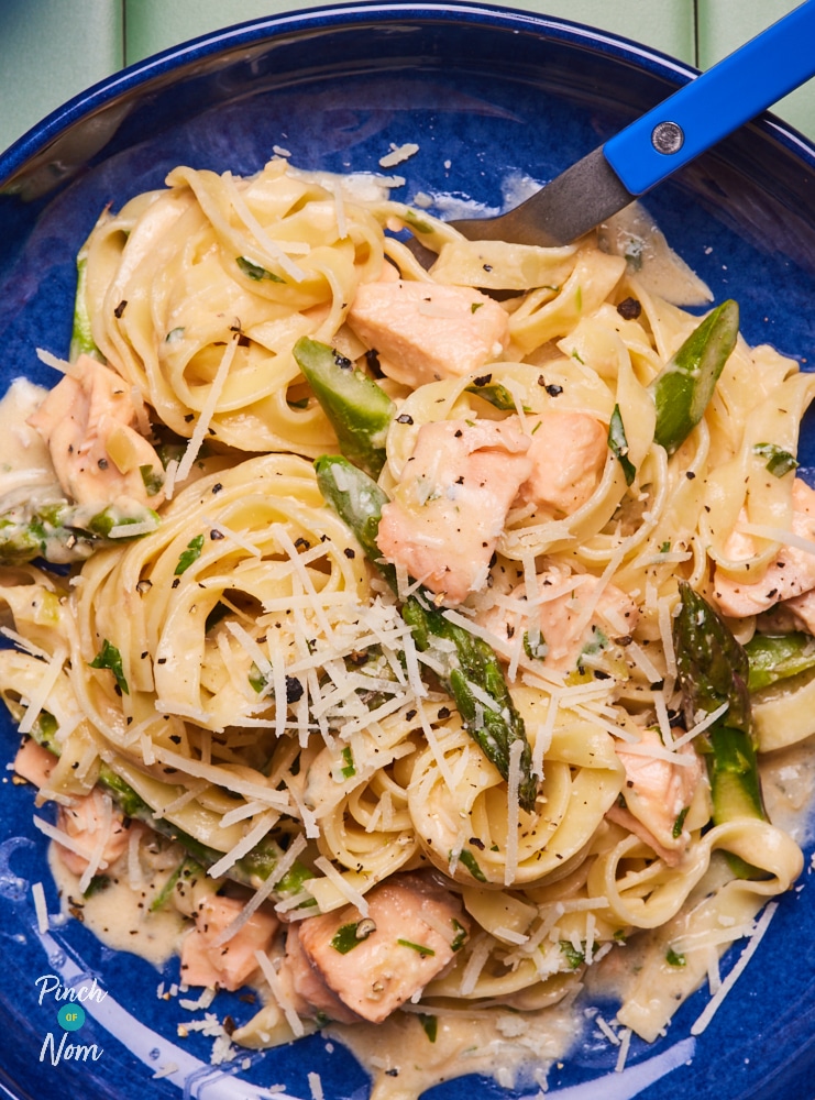 Pinch of Nom's slimming-friendly Salmon and Asparagus Pasta is served on a deep blue plate, while a matching fork digs in.