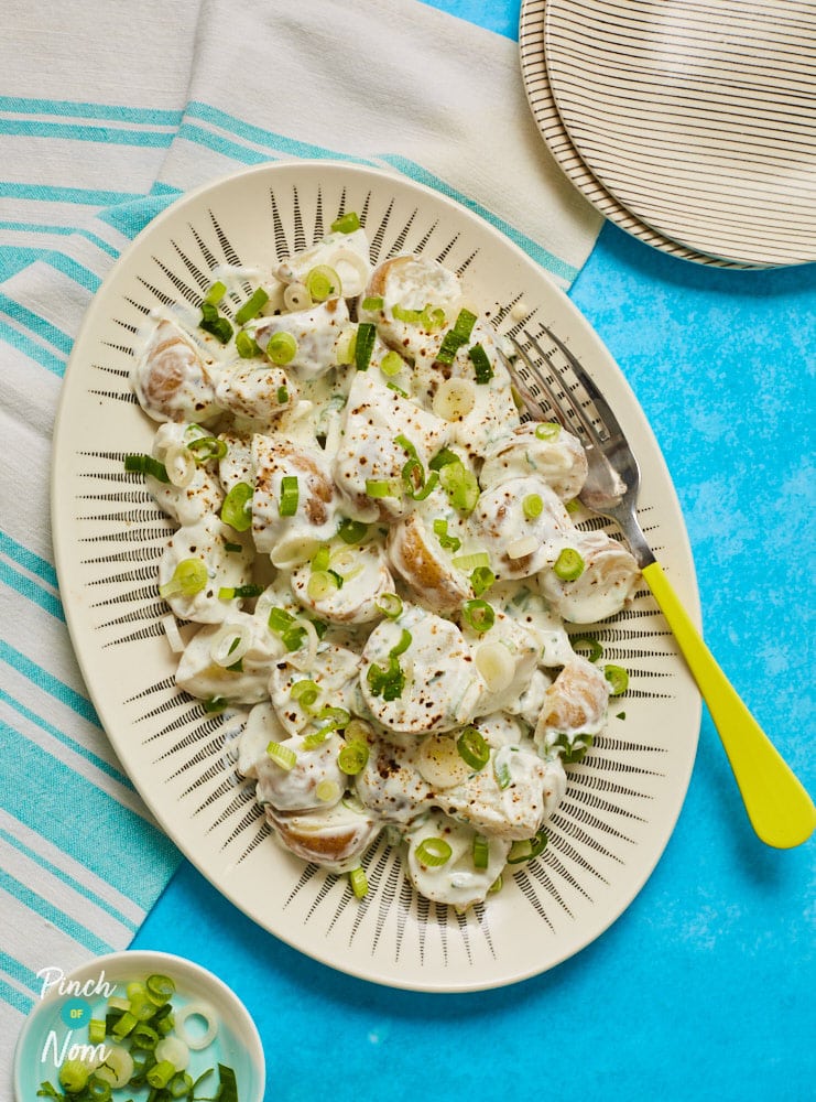 Pinch of Nom's Potato Salad is served on a large plate, coated in a creamy sauce with plenty of spring onion and chives stirred through. A fork is waiting to tuck in, with side plates ready to serve.