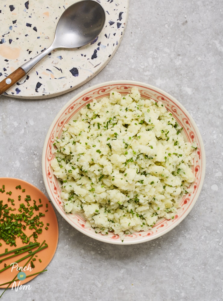 Pinch of Nom's Creamy Mashed Potatoes are served in an orange and white bowl with chopped-up chives and spring onions stirred through.