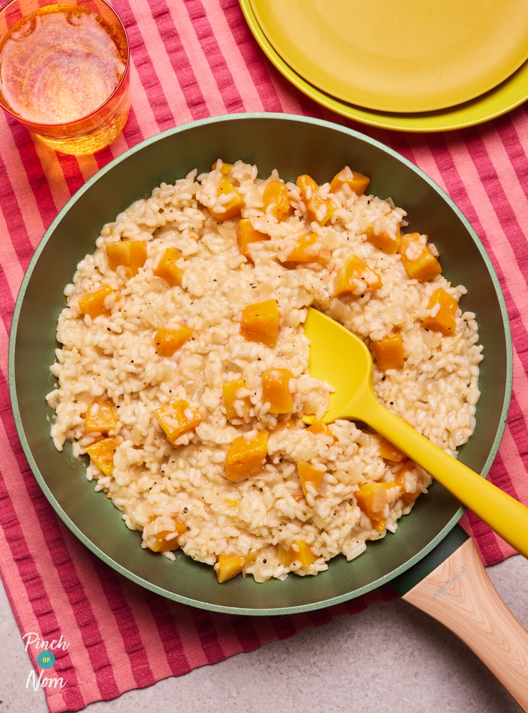 Pinch of Nom's Butternut Squash Risotto is served in a pan, set on a red and pink striped cloth. The risotto is creamy, with chunks of orange butternut squash. A yellow serving spoon rests in the pan, ready to dish up.
