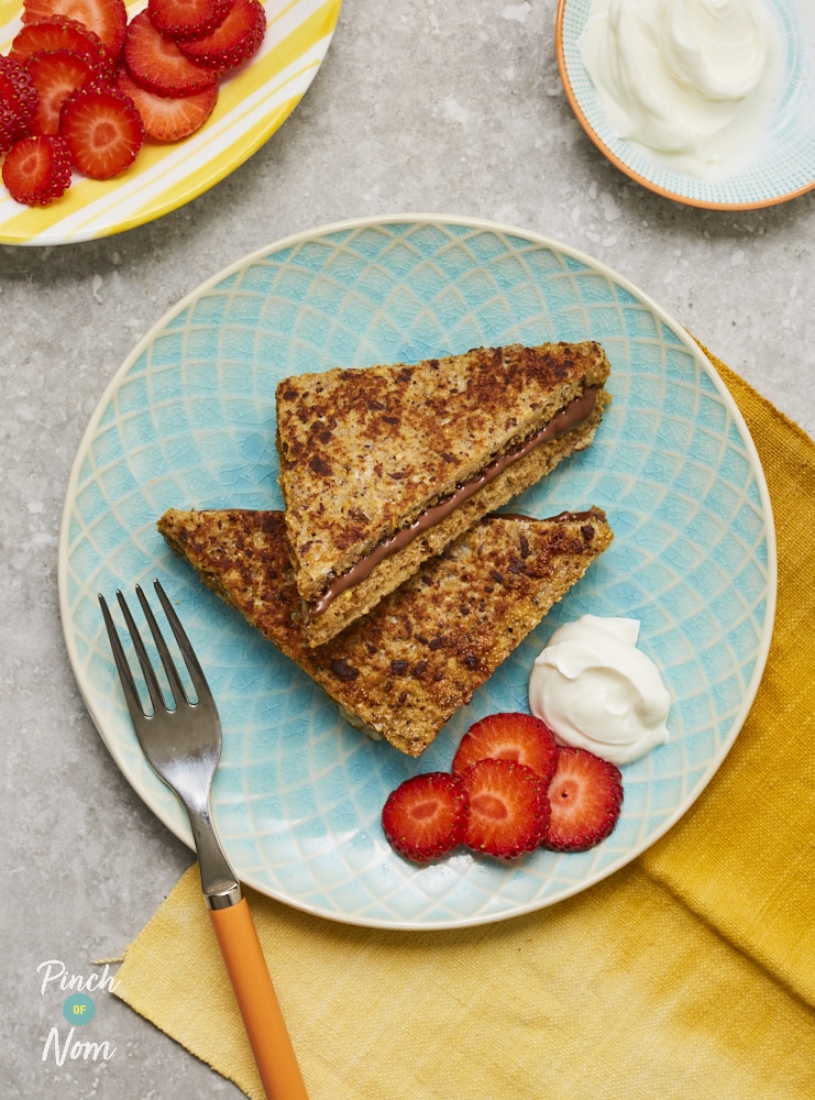 Pinch of Nom's Chocolate French Toast is served on a summery blue plate with a swirl of aerosol cream and juicy, sliced strawberries. There are plates to the side with extra sliced strawberries and cream, and a yellow fork waiting to tuck in.