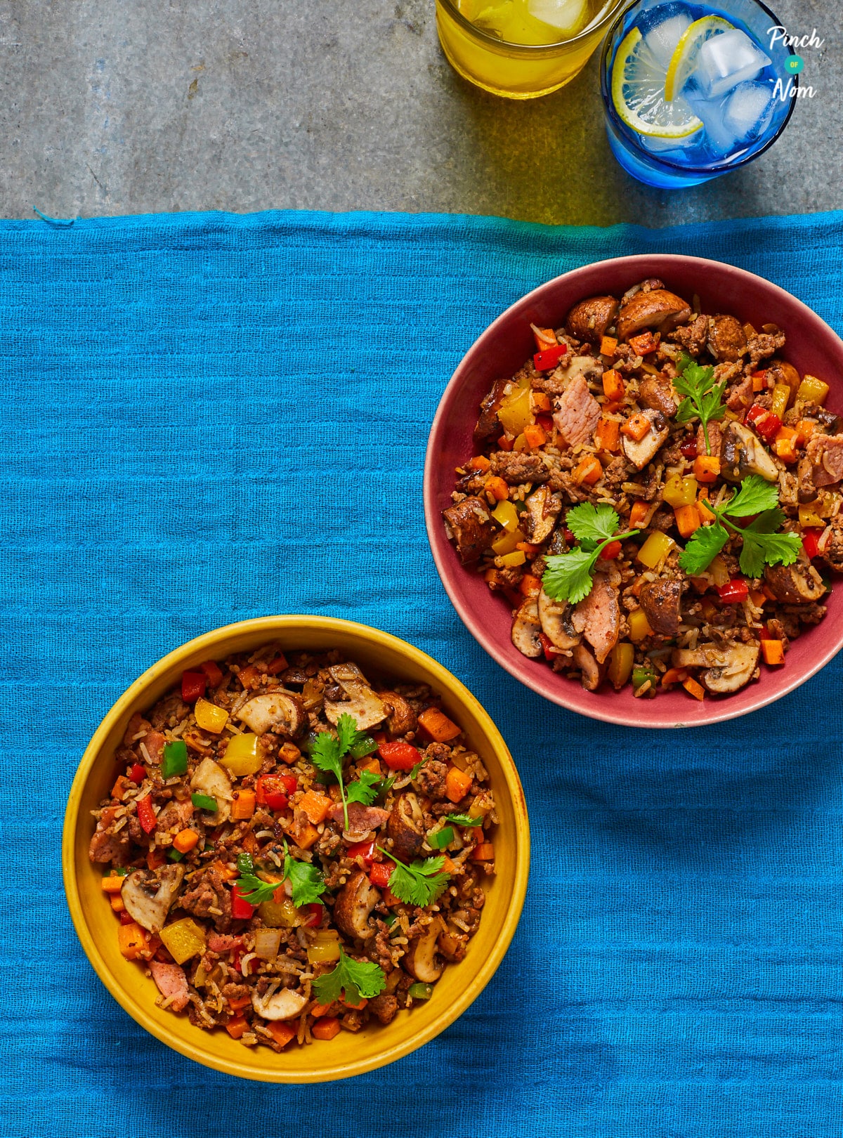 A table is set with a blue tablecloth, two glasses of water with ice cubes and slices of lemon, and two bowls of Pinch of Nom's slimming-friendly Cajun-Style Dirty Rice. The rice is a medley of colourful vegetables including carrots, peppers and mushrooms, topped with a sprig of fresh green coriander.