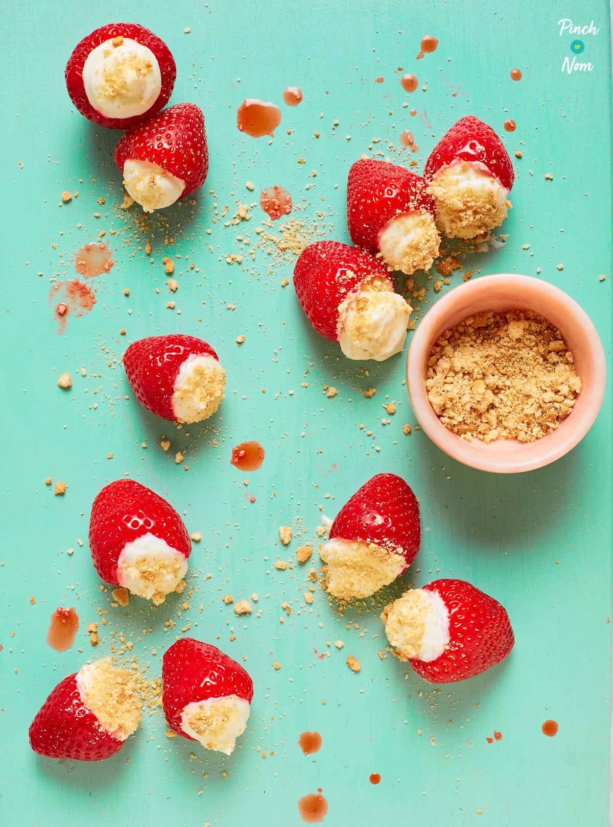 Pinch of Nom's Cheesecake Stuffed Strawberries are scatted on a turquoise surface, dusted with biscuit crumbs. A small pink pot is nearby, filled with crushed biscuit. The tops of the strawberries have been cut off, and a cheesecake-style filling has been piped into each berry.
