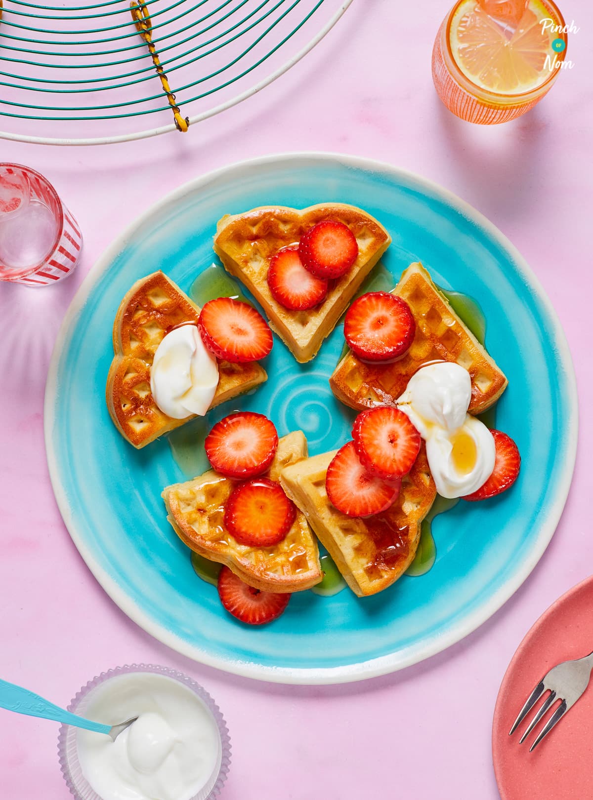 A table is set with a plate full of Pinch of Nom's slimming-friendly strawberry and vanilla waffles, topped with low-sugar syrup sliced strawberries and a dollop of sweetened yoghurt.