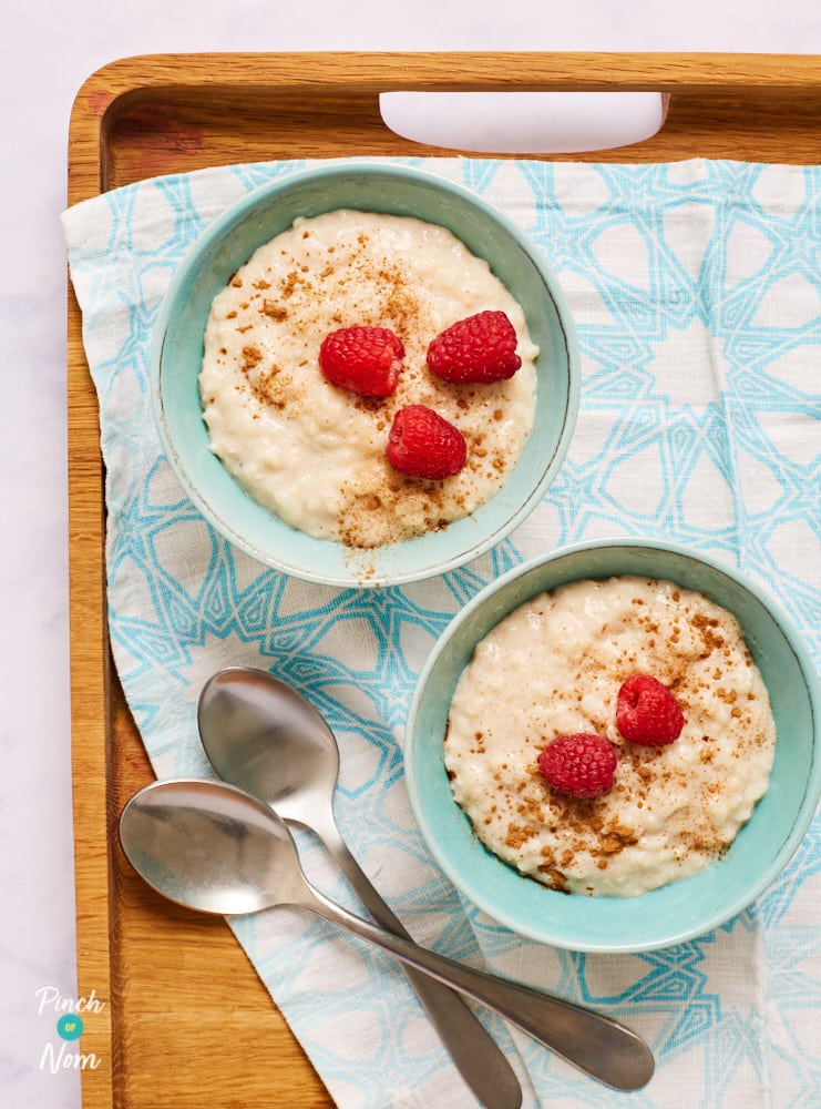 Two bowls of Pinch of Nom's slimming-friendly Chai Rice Pudding are served on a tray, topped with berries and a sprinkling of cinnamon, with two spoons waiting to tuck in.