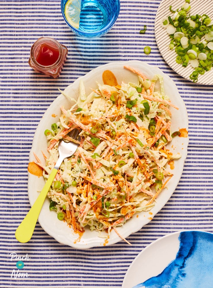 Pinch of Nom's slimming-friendly Buffalo Coleslaw is ready to eat, served on a large white serving platter. The platter sits on a table, covered with a stripy tablecloth. A yellow-handled fork is placed on top of the coleslaw, ready to scoop up the finely shredded vegetables.