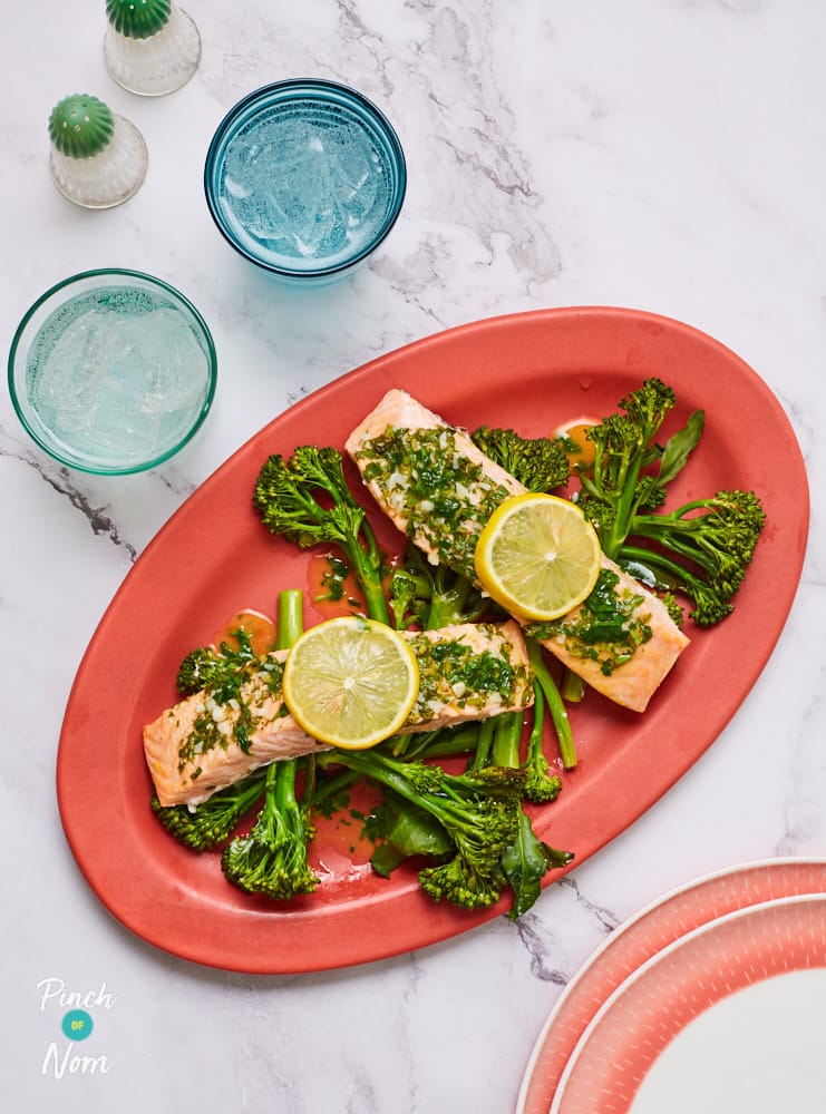 2 fillets of Pinch of Nom's Baked Garlic Salmon are served on a large, red serving plate on a bed of tenderstem broccoli with a garnish of lemon wedges.