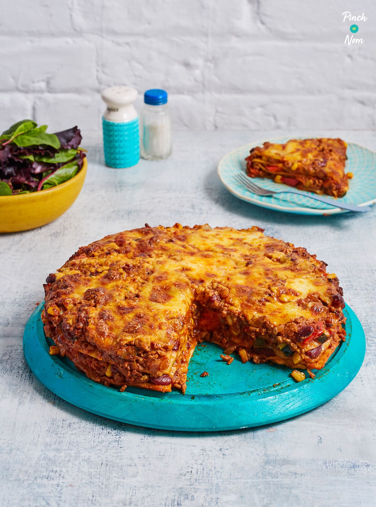 Pinch of Nom's slimming-friendly Enchilada Lasagne is served at the centre of a table with a large slice removed, revealing the layers inside.