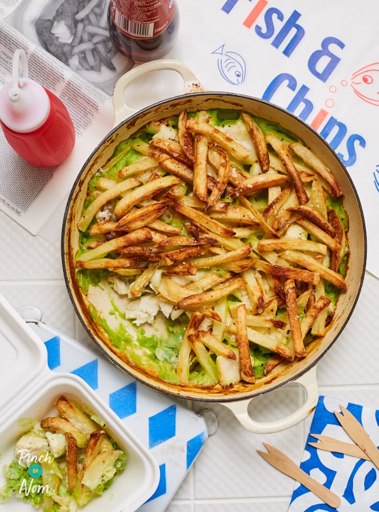 Pinch of Nom's Fish and Chip Pie is served in a large, round casserole dish. Underneath a topping of crispy, golden chips, you can see flaky white fish and vibrant green mushy peas. A squeezy bottle of ketchup is nearby, and a portion of the recipe has been spooned into a takeaway style polystyrene carton. 