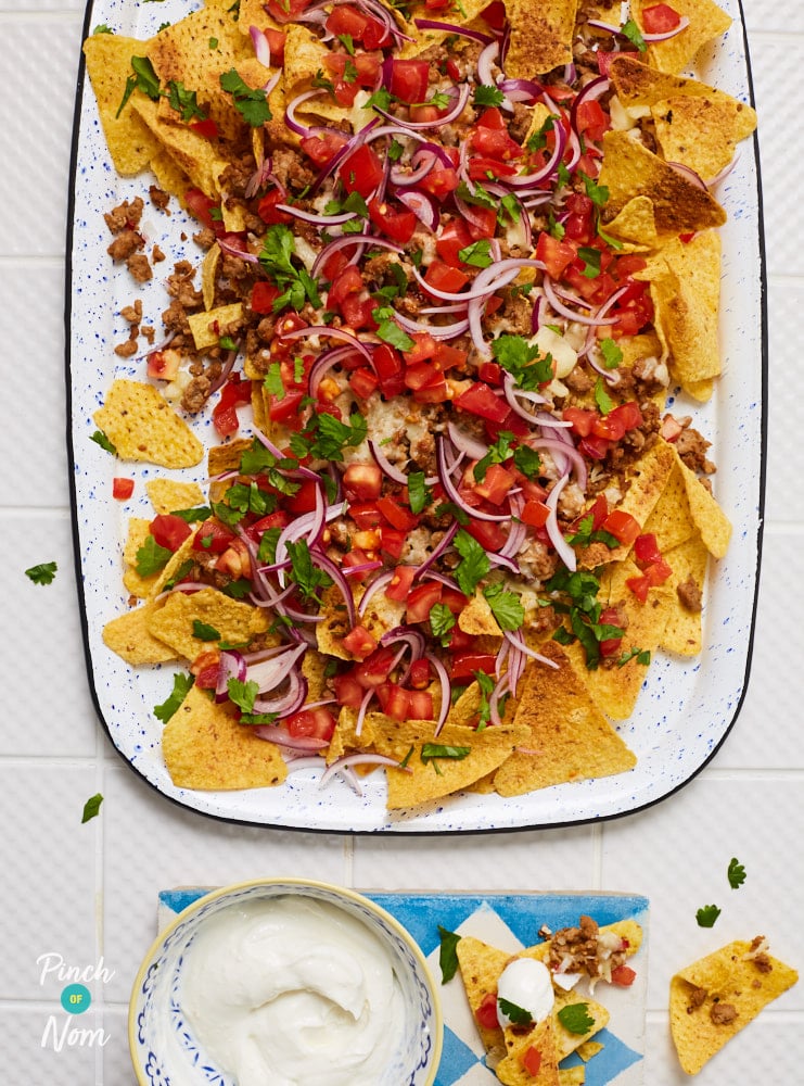 Pinch of Nom's colourful Honey and Lime Pork Nachos are served in a large dish at the centre of a table with a homemade mix of garlic-infused yoghurt in a small bowl to the side, waiting to be dipped.