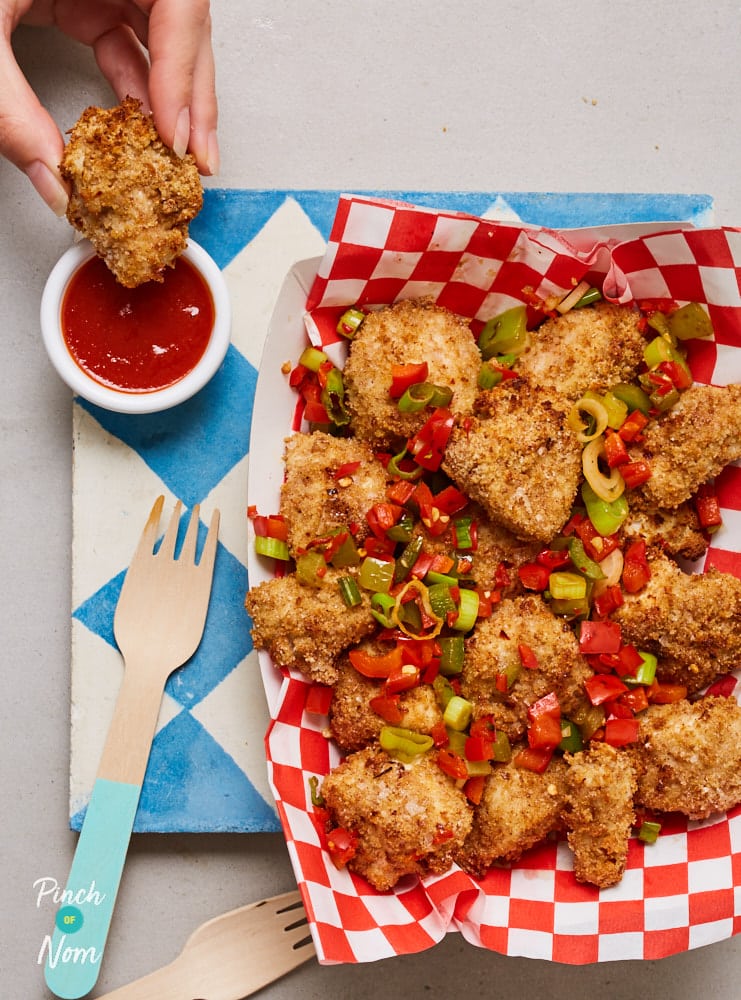 A street food style table is set with Pinch of Nom's Salt and Pepper Chicken Nuggets served on red and white striped paper, with dipping sauce to the side, and a wooden fork waiting to tuck in.