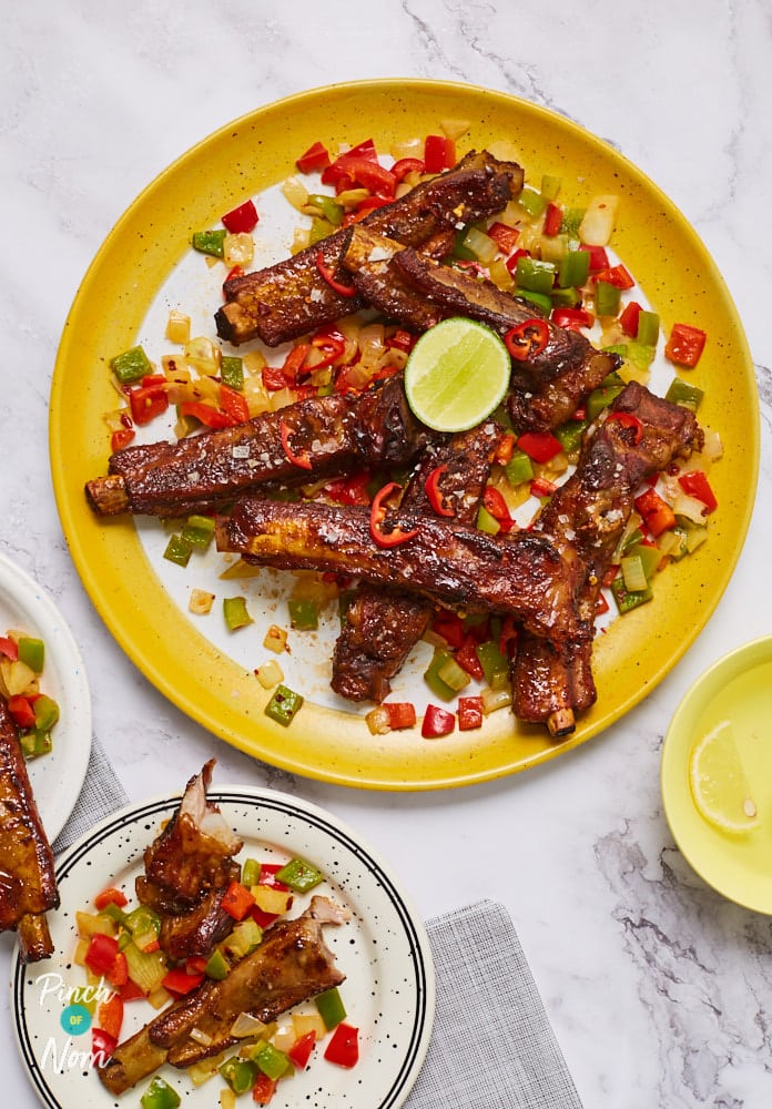 A white marble table is set with a large yellow plate of Pinch of Nom's Salt and Pepper Ribs. The ribs look juicy and golden, served with chopped peppers and onions, and garnished with a slice of fresh lime. Two smaller plates are also on the table, and a portion of ribs has been served onto each one.