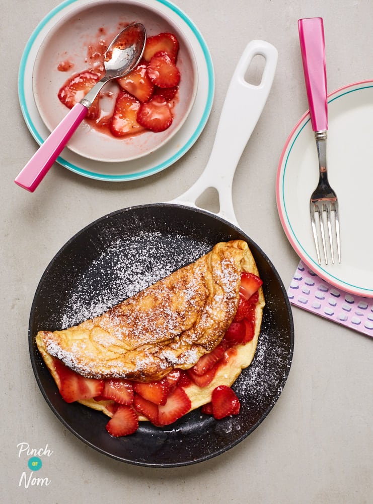 Pinch of Nom's Strawberry Souffle Omelette is ready to serve inside a small frying pan; it's dusted with icing sugar while the sliced strawberry filling spills out. There are extra sliced, juicy-looking strawberries waiting on a side plate with a fork.