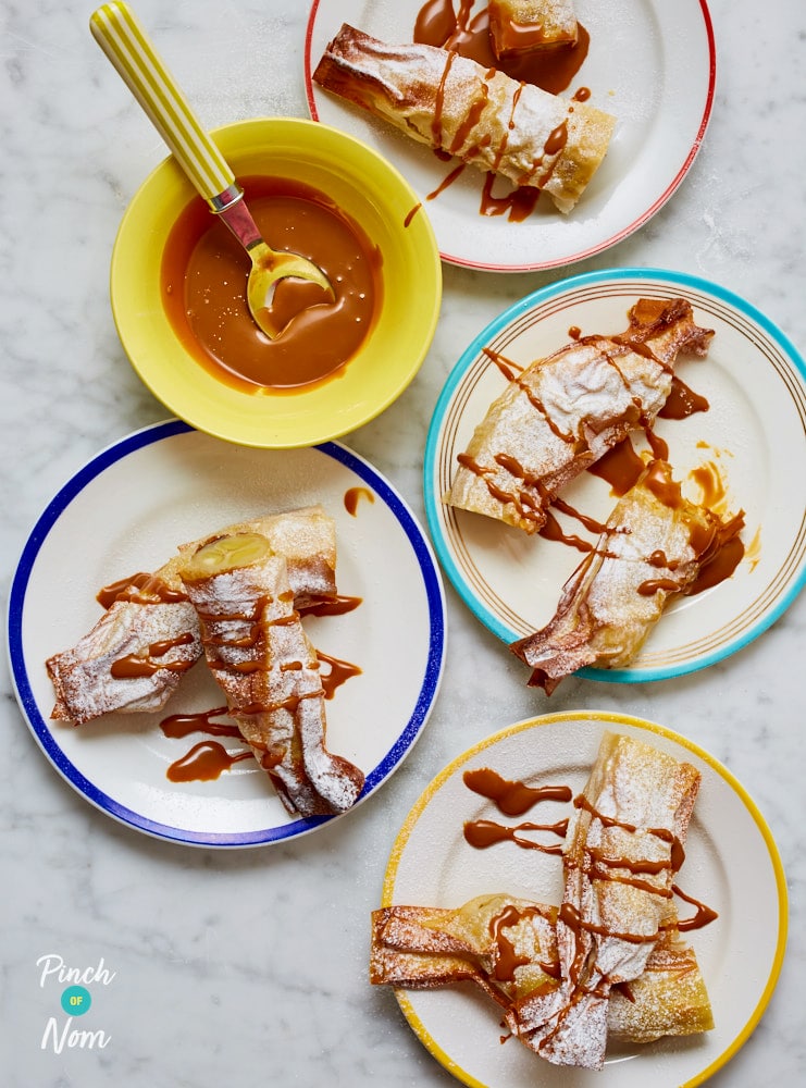 A table is set with four plates of Pinch of Nom's Banana and Caramel Filo Rolls. They're all ready-decorated with icing sugar and a caramel drizzle, but a spoon waits to add more on top from a small bowl set to the side.