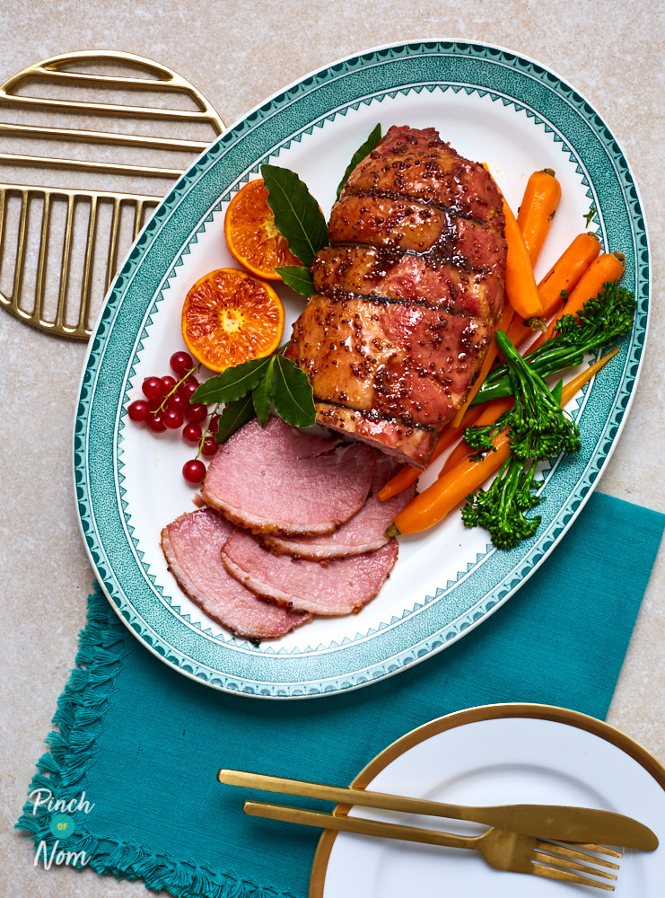 Pinch of Nom's slimming-friendly Ginger and Orange Glazed Ham is served over a bad of roast veggies; the joint is partially carved and ready to serve.