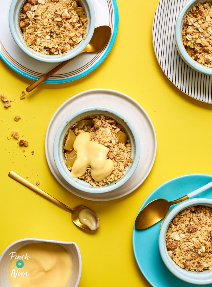 Four portions of Pinch of Nom's Pear and Ginger Crumble served in individual ramekin dishes on a yellow tabletop, with a jug of custard to the side waiting to pour over.