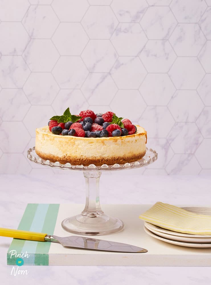Pinch of Nom's slimming-friendly Raspberry and Blueberry Baked Cheesecake is served at the centre of a table decorated with berries and icing sugar.