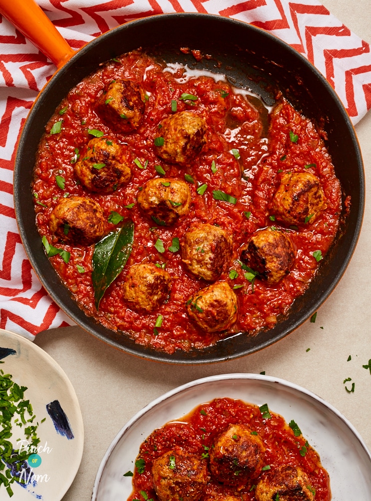 A large pan is filled with freshly-made Spanish-Style Meatballs. Chopped, flatleaf parsley pokes through the colourful red sauce. The meatballs are browned and juicy.