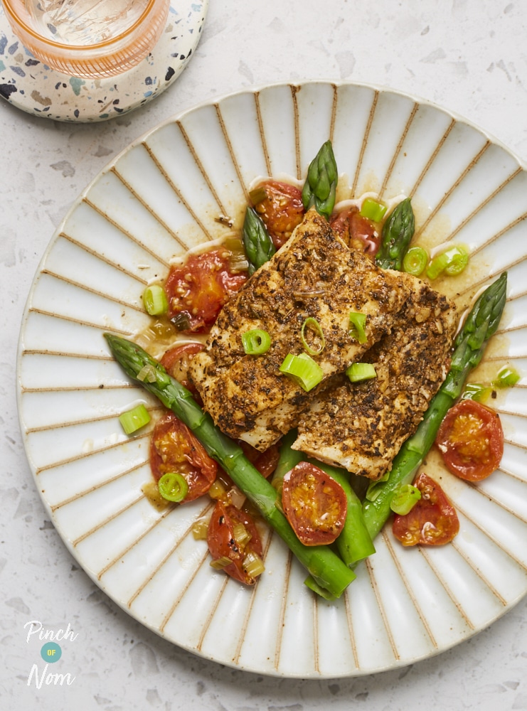 A table is set with a plate of Pinch of Nom's Cajun Baked Cod with Asparagus and Cherry Tomatoes. The cod fillets are served on a colourful bed of vegetables, and a sprinkling of spring onions on top.