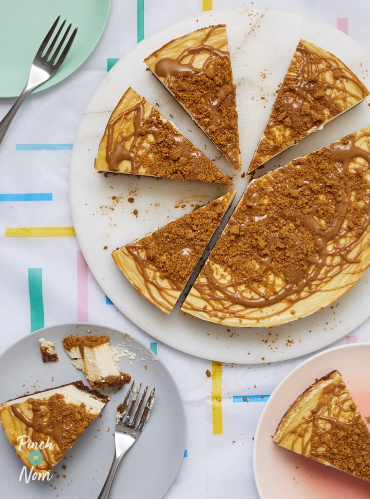 Pinch of Nom's slimming-friendly Biscoff Cheesecake is served on a large plate at the centre of a table; half has already been sliced, with a slice served on two separate side plates.
