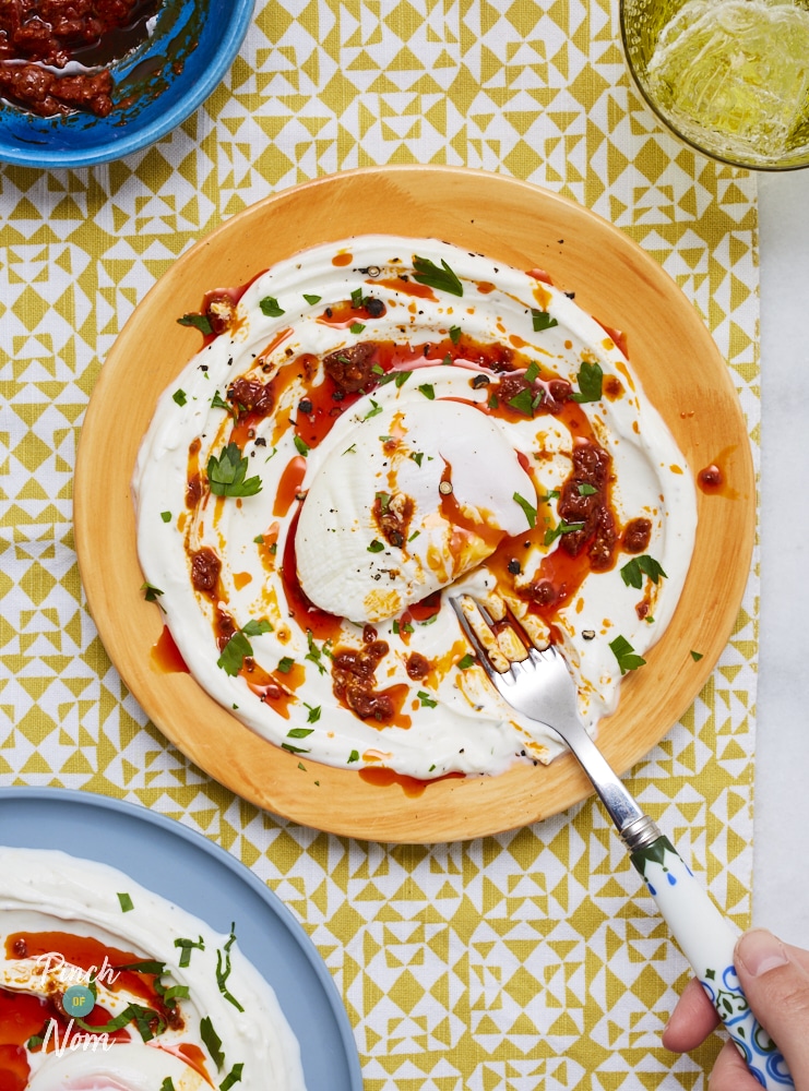 Pinch of Nom's Turkish Style Eggs are plated up on summer tableware. The plate is swirled with yoghurt and harissa and topped with egg, while a fork waits to tuck in.