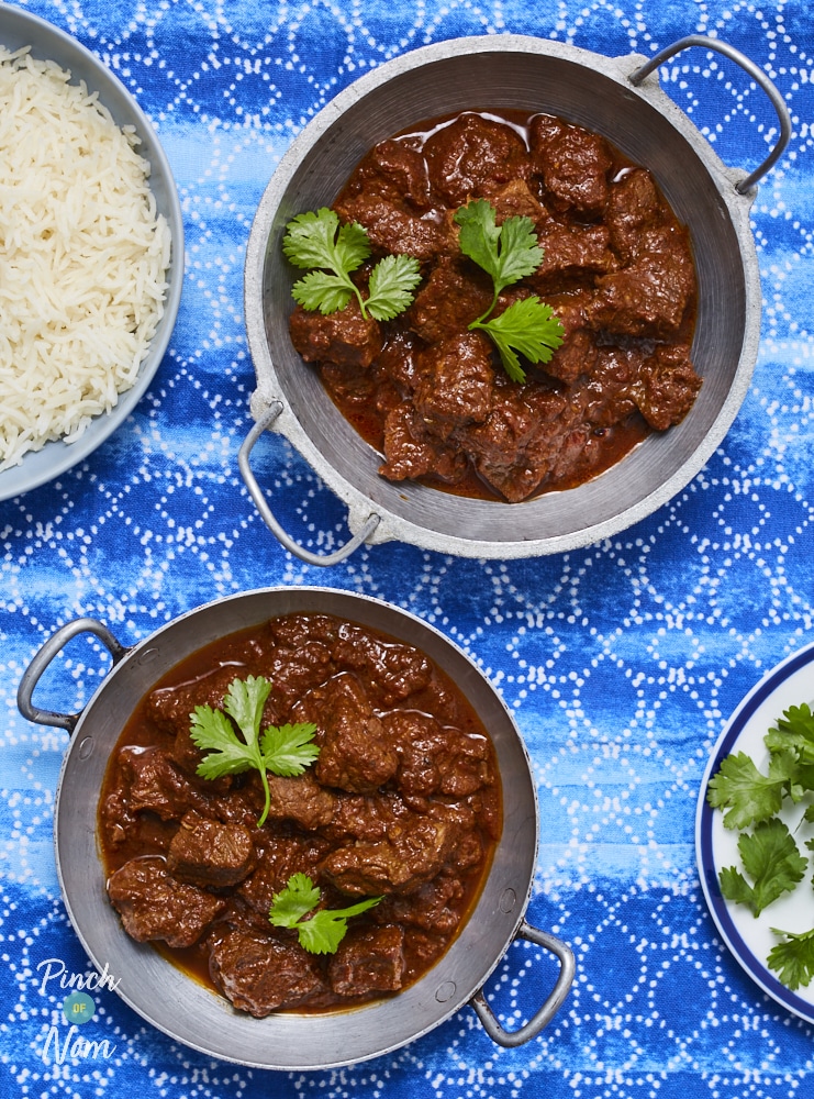 Two silver bowls are filled with Pinch of Nom's Beef Madras, set on a blue patterned tablecloth. Each bowl is garnished with fresh green coriander and a bowl of rice is nearby for serving.