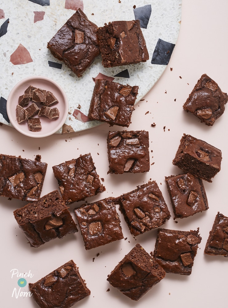 A batch of Pinch of Nom's Chocolate Orange Brownies is served on a bright pink tabletop, spilling from a speckled serving dish. The squares look gooey and squidgy in the middle with chocolate chips poking through.