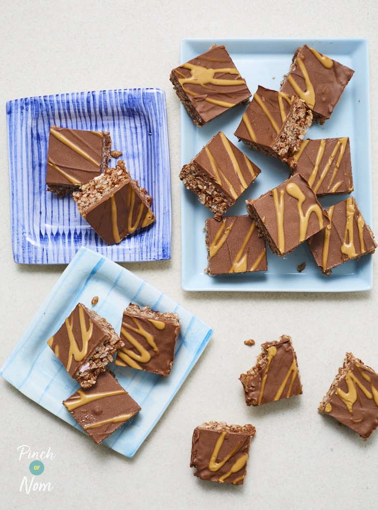 Pinch of Nom's Chocolate Peanut Butter Bars are served in three batches on three different square, blue serving plates. They're all topped with a low-calorie peanut drizzle and one has already been bitten.