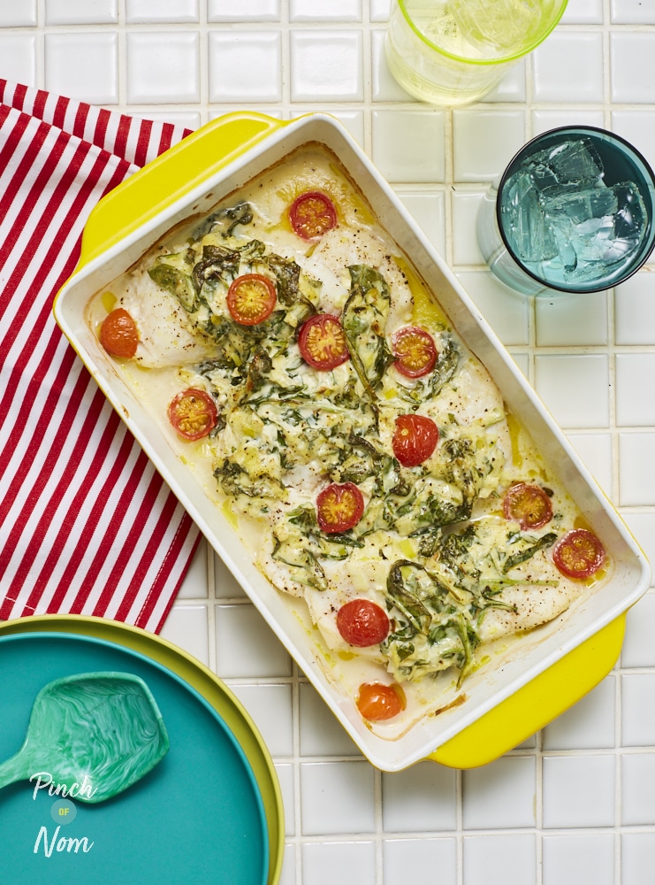 Pinch of Nom's Creamy Cod and Spinach is served in a large, rectangular oven dish. The dish sits on top of a white, tiled countertop, with a red and white striped tea towel underneath. The flaky fish is coated in a creamy spinach sauce, with cherry tomatoes scattered on the top. 