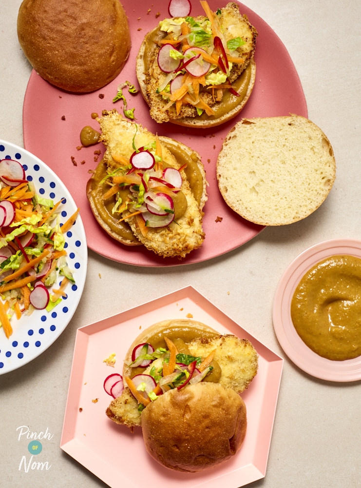 Pinch of Nom's Katsu Curry Burgers spilling onto pink hexagonal serving plates; the burger buns are open to reveal the crispy chicken patties inside, covered in a drizzle of Katsu-style sauce and fresh colourful salad veg.