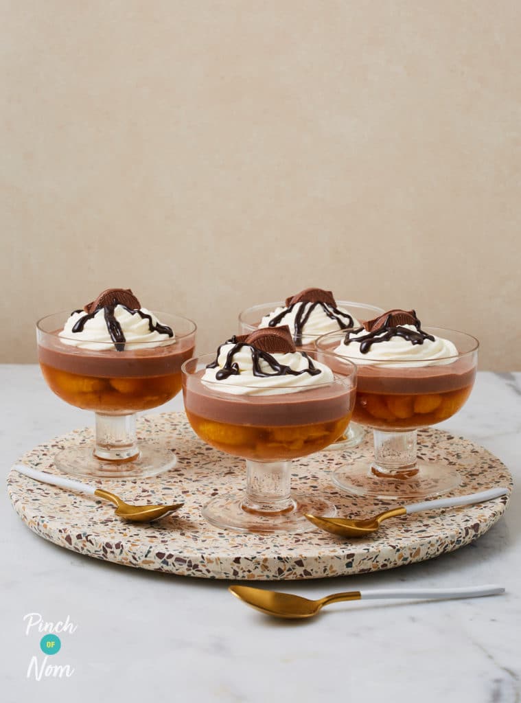 Four of Pinch of Nom's Chocolate Orange Trifles are served in glass trifle dishes on a round serving board. There are spoons to the side waiting to tuck in. Each trifle is topped with a drizzle of chocolate sauce and Terry's Chocolate Orange segments.