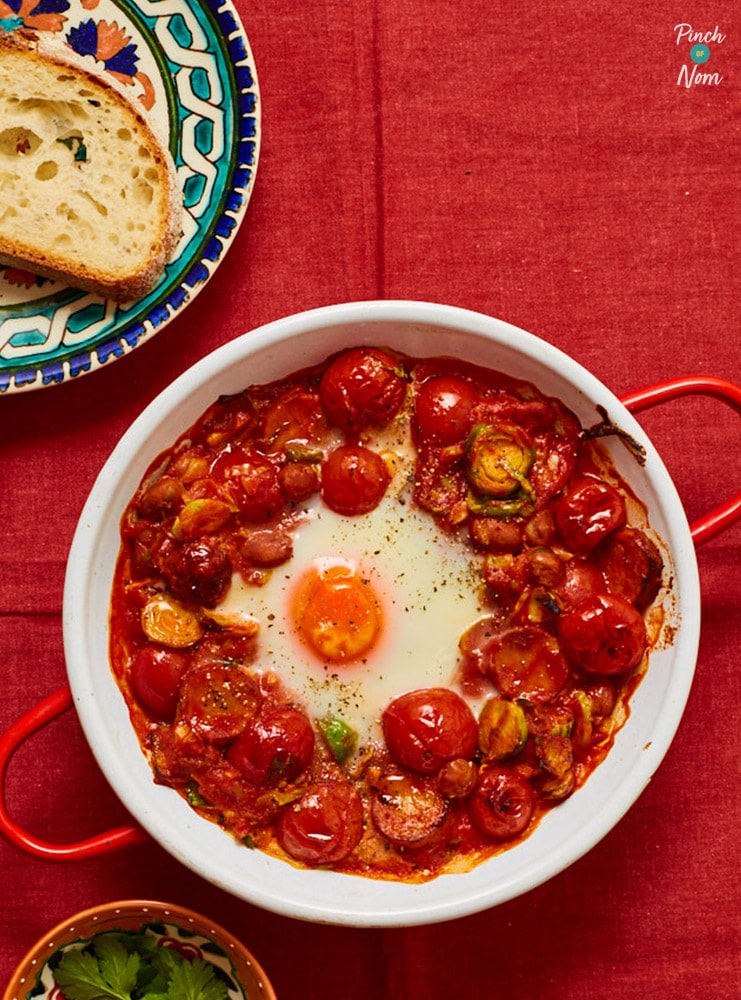 A portion of Pinch of Nom's Spanish Eggs is served in a red ovenproof dish with crusty wholemeal sliced bread to the side, waiting to be dipped in. The whole portion is topped with a sunny-side-up fried egg.