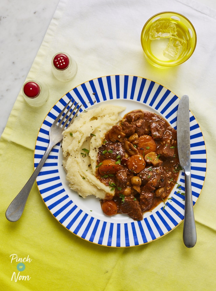 A blue and white striped plate is set on top of a pale yellow table cloth. The plate is filled with Pinch of Nom's Beef Bourguignon recipe, and a heap of Creamy Mashed Potatoes. A silver knife and fork are laid on top of the plate, ready to dig in.