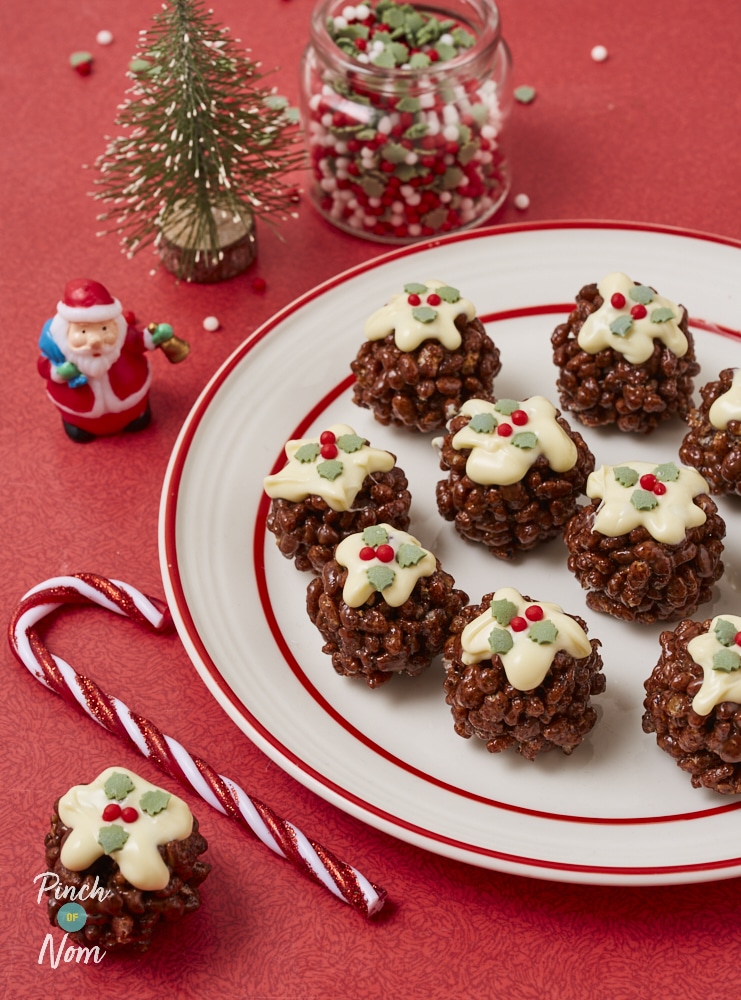 A large plate is filled with a batch of Pinch of Nom's slimming-friendly Rice Krispie Christmas Puds, served on a bright red festive table, decorated with candy canes and mini Christmas trees.