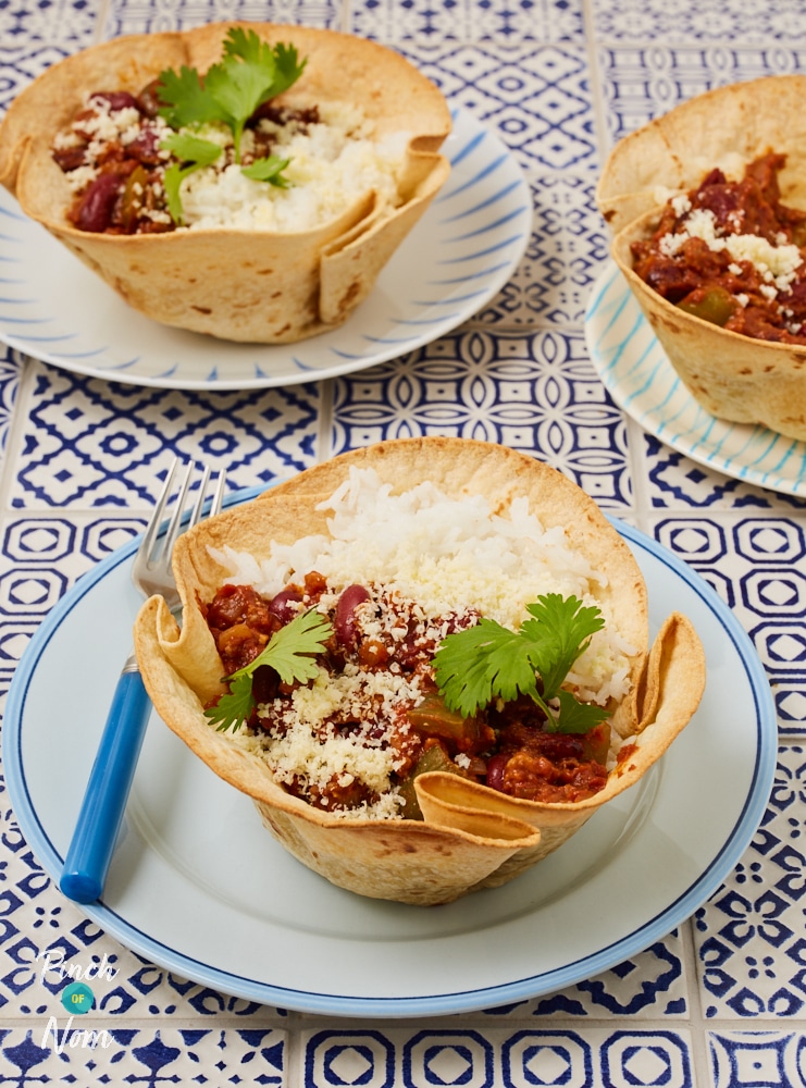 Three plates are set on a tiled countertop. Each plate is filled with a crisp, golden tortilla wrap that has been baked into a bowl shape. Inside the tortilla bowls are fluffy white rice and rich brown chilli con carne, sprinkled with cheese.
