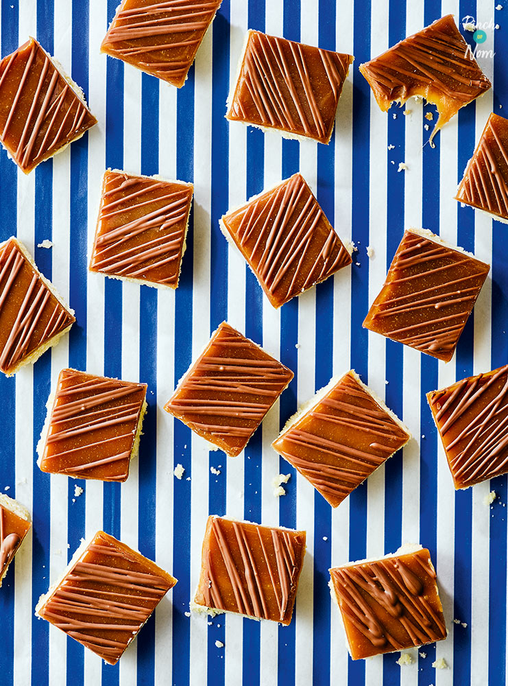 Squares of Pinch of Nom's Millionaire's Shortbread are drizzled with a sticky caramel topping, and scattered across a blue and white striped tabletop.