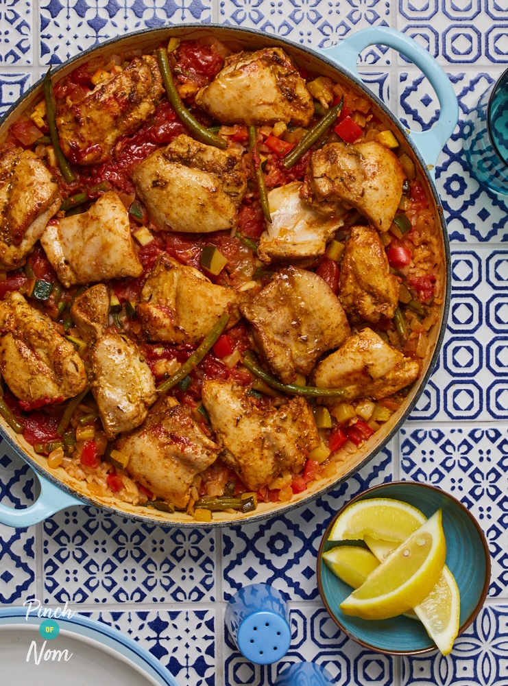 A large blue dish is filled with Pinch of Nom's slimming-friendly One-Pot Chicken and Tomato Rice on a tiled mosaic-patterned surface; there are lemon wedges to the side for squeezing on top before serving.