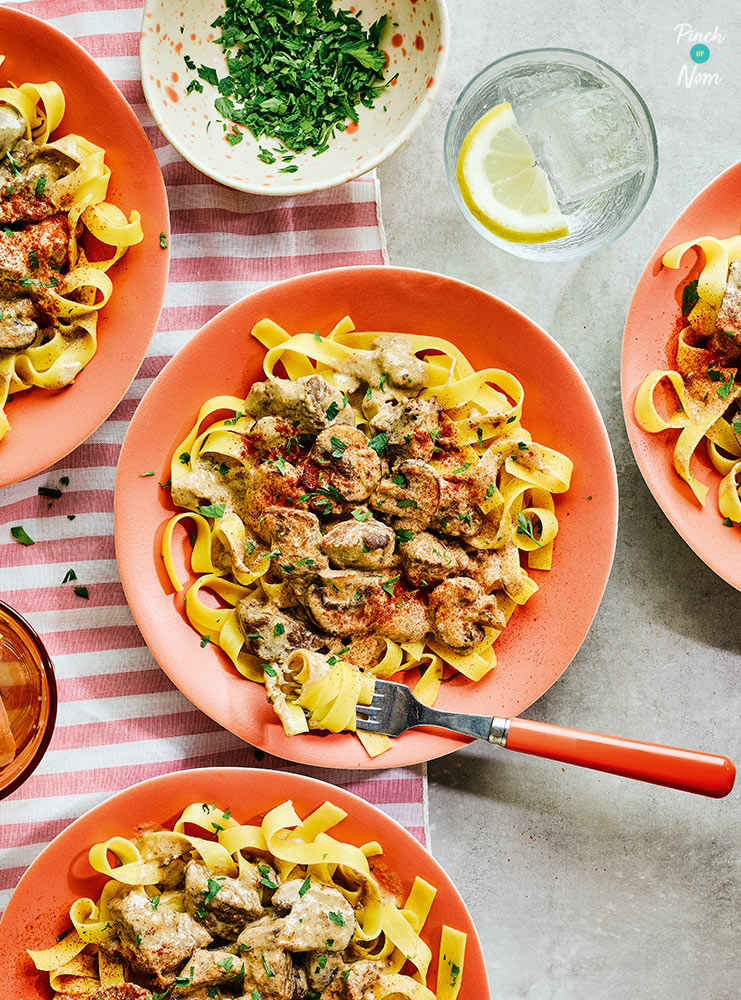 A table is set with four orange plates. Each plate is filled with Pinch of Nom's Slow Cooker Stroganoff. Creamy beef and mushrooms are served on a bed of tagliatelle pasta, dusted with paprika and a sprinkling of fresh green herbs.
