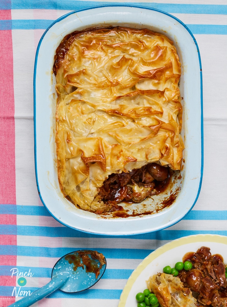 An oven dish is filled with a golden-topped portion of Pinch of Nom's slimming-friendly Steak and Stilton Pie; a portion has been spooned onto a plate to the side, revealing the meaty middle.