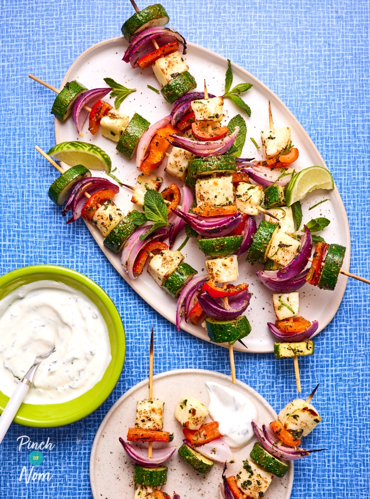 A large serving plate is filled with a batch of Pinch of Nom's slimming-friendly Minty Halloumi and Vegetable Kebabs, with a fresh yoghurt dipping sauce to the side.